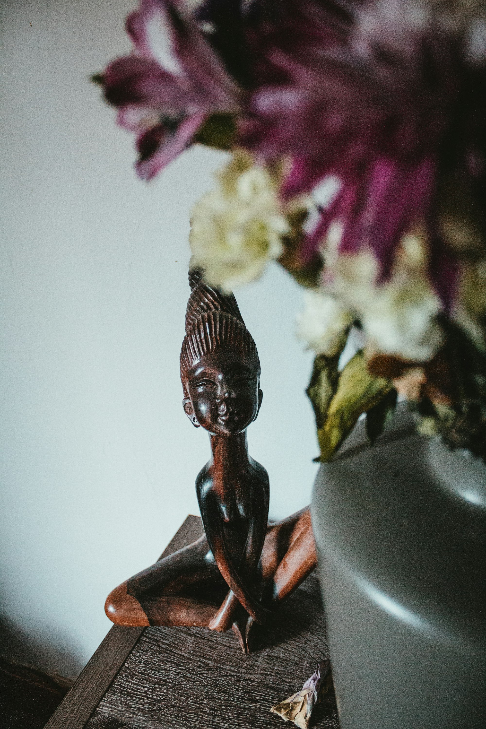 A dark teak Balinese sculpture beside a vase of tropical blooms on a timber surface.
