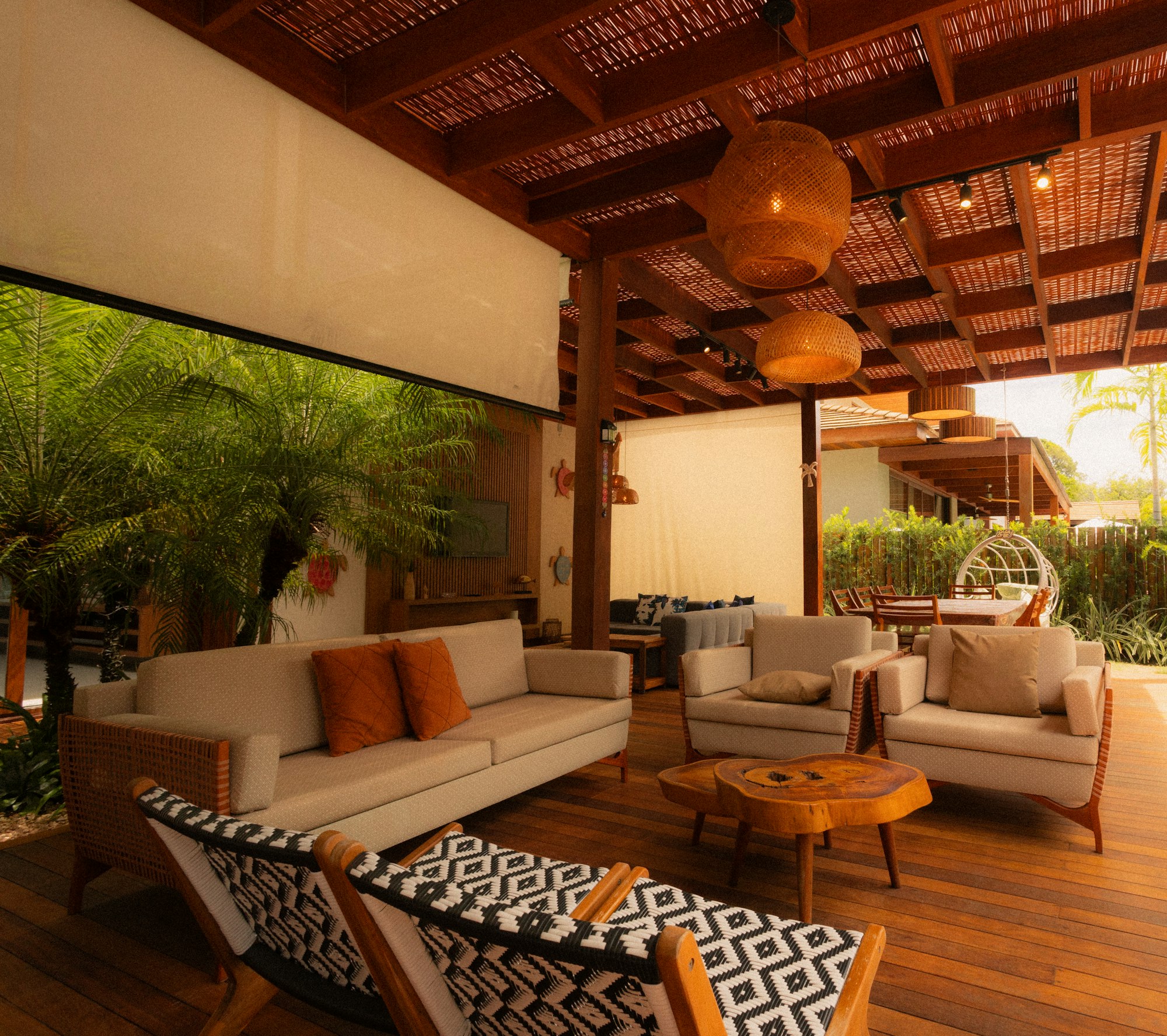 An open-air tropical lounge with a woven-roof pergola, rattan pendant lights, and lush planting.