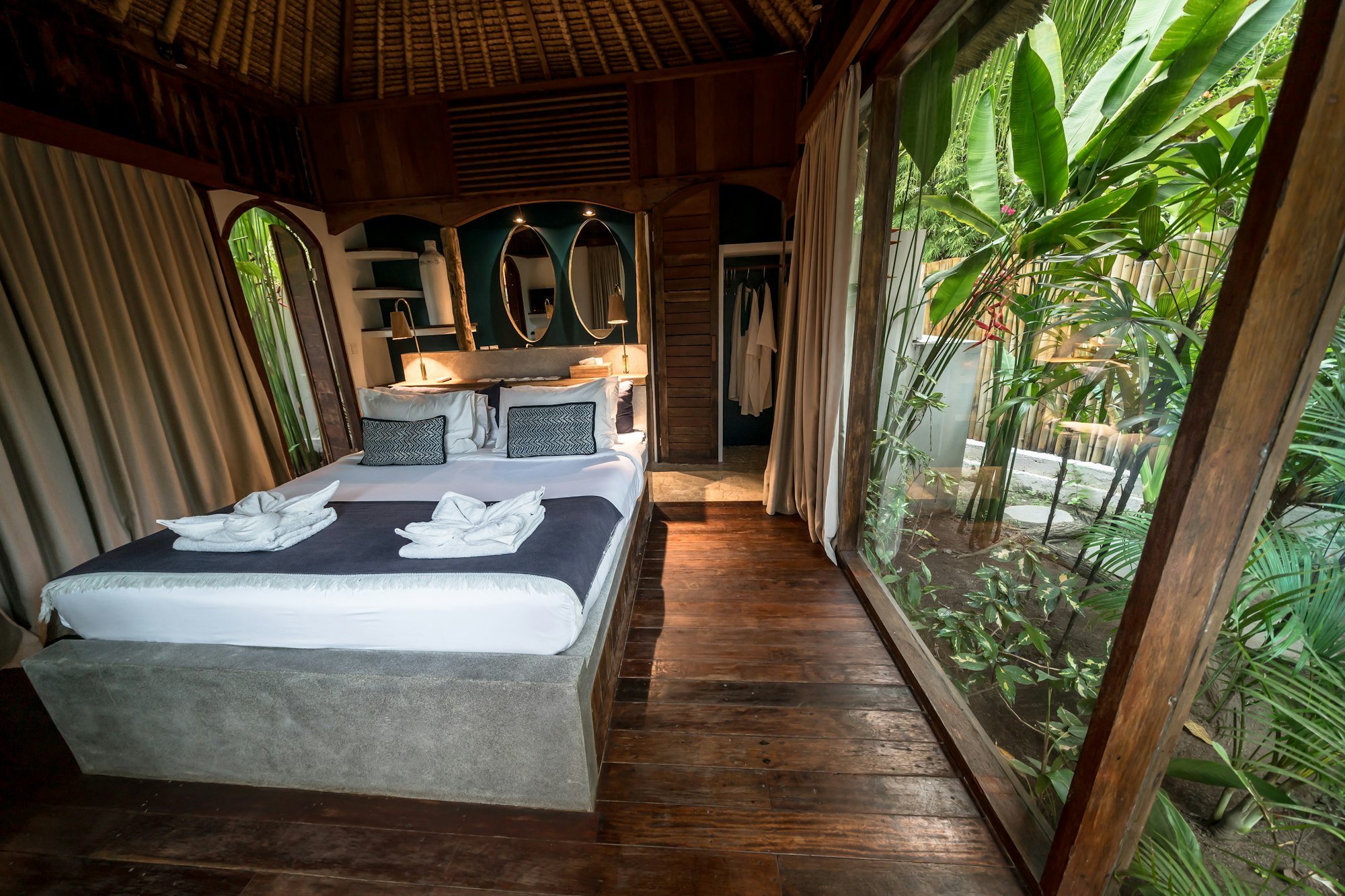 A luxury tropical bedroom with a low platform bed, exposed wooden beams, and floor-to-ceiling glass opening onto a lush garden.