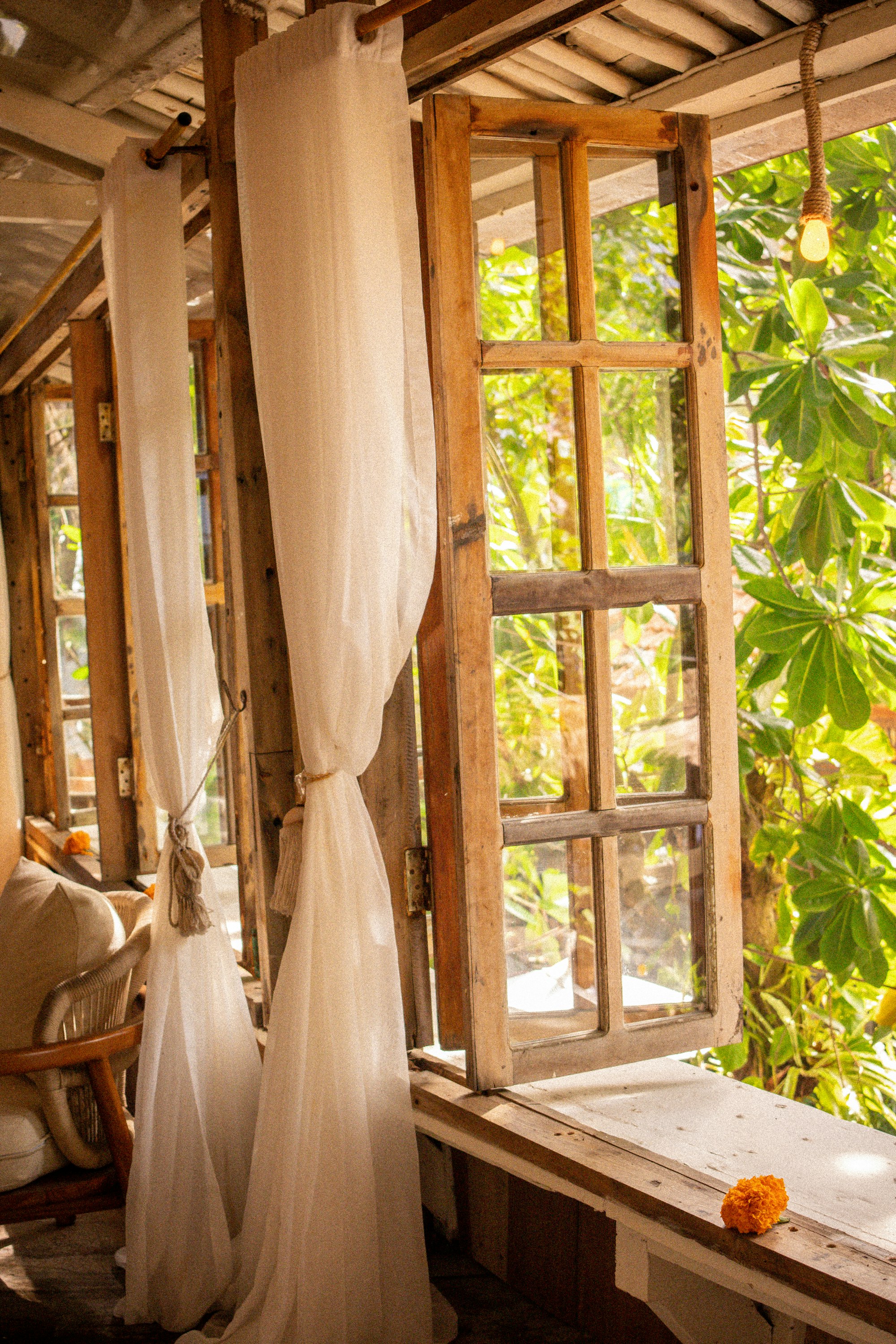 A sunlit tropical bedroom nook with a sheer linen curtain, an open wooden window, and a rattan chair overlooking greenery.